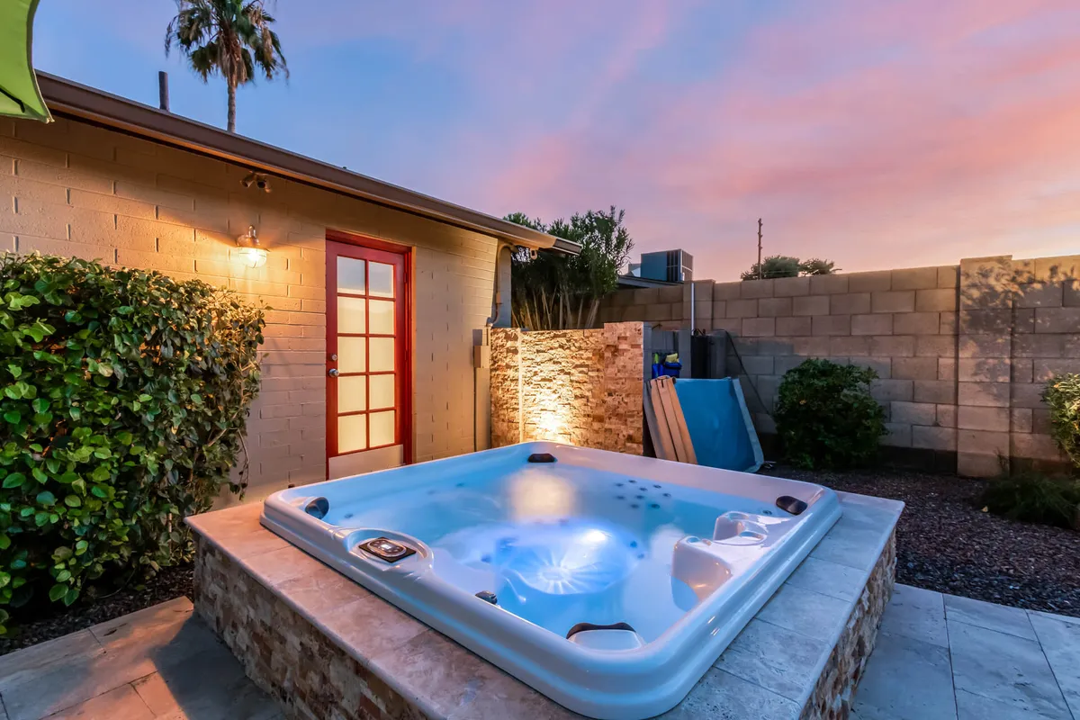 An inviting hot tub set against a backdrop of lush landscaping, a bold red door, and desert sunset views. Social seating and warm lighting evoke a sense of evening relaxation and camaraderie.