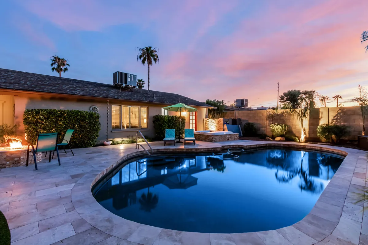 A serene backyard scene with a sparkling swimming pool, several lounge chairs arranged for sunbathing, a cozy fire pit, and tall palm trees swaying above lush greenery. The space is designed for both relaxation and social gatherings.