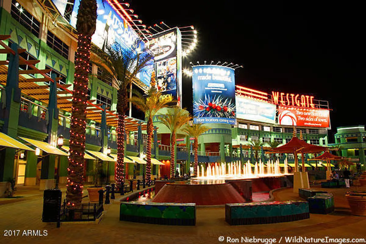 A lively scene of a public plaza with a central fountain, tall palm trees, neon signs, and colorful buildings. People stroll and gather, highlighting the area's festive, community-oriented ambiance.