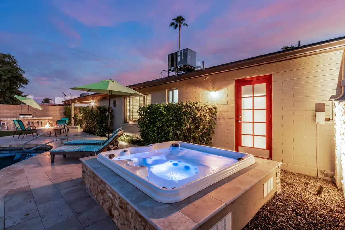 A vibrant backyard view showcasing a hot tub adjacent to a swimming pool, with green umbrellas offering shade, and a fire pit for evening gatherings. The arrangement of patio furniture invites group relaxation and socializing.