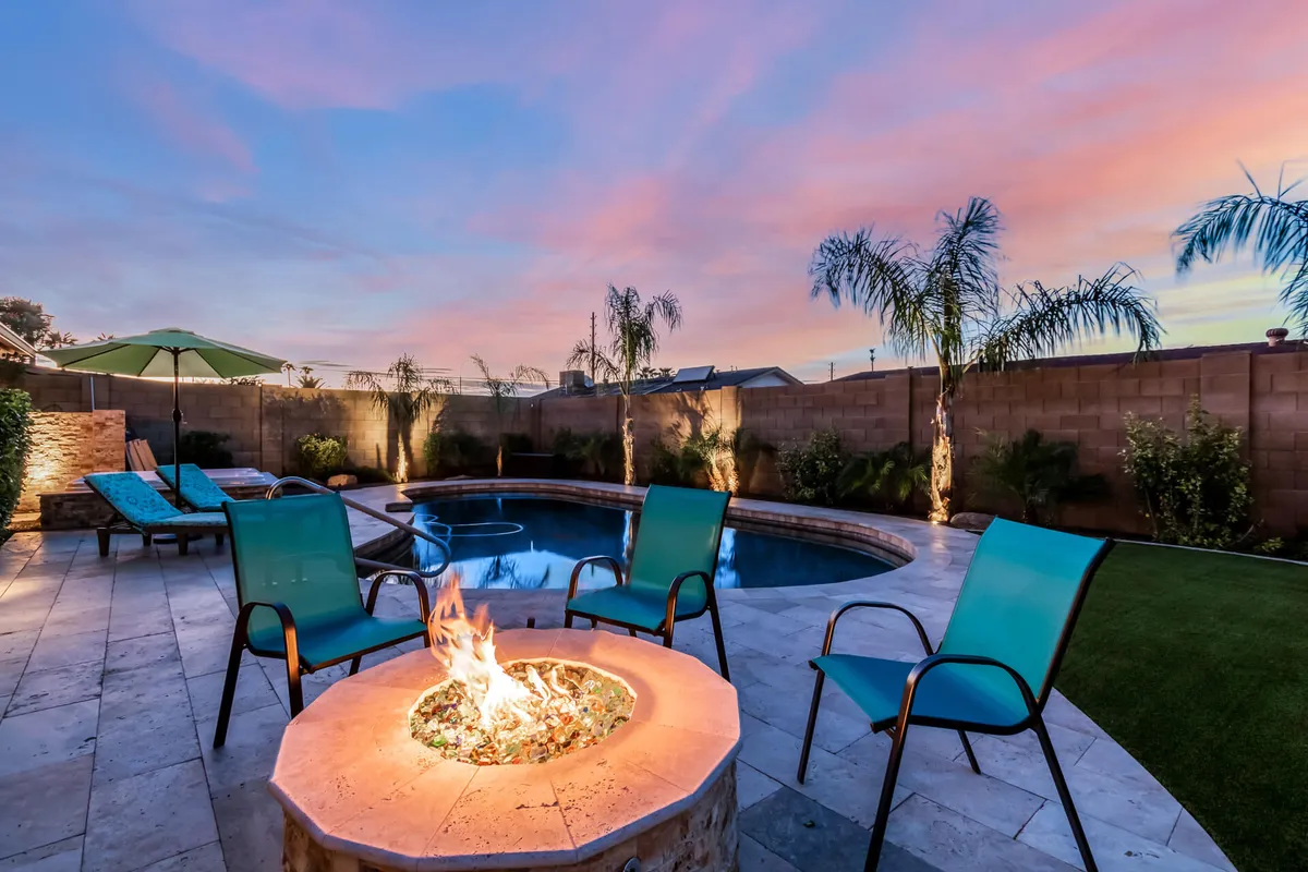 The image showcases the backyard oasis of a Glendale, Arizona single-family home, featuring a sparkling swimming pool surrounded by lush palm trees, inviting lounge chairs under an umbrella, and a cozy fire pit area perfect for evening gatherings. The scene is bathed in the warm glow of sunset, creating a relaxed and welcoming atmosphere.