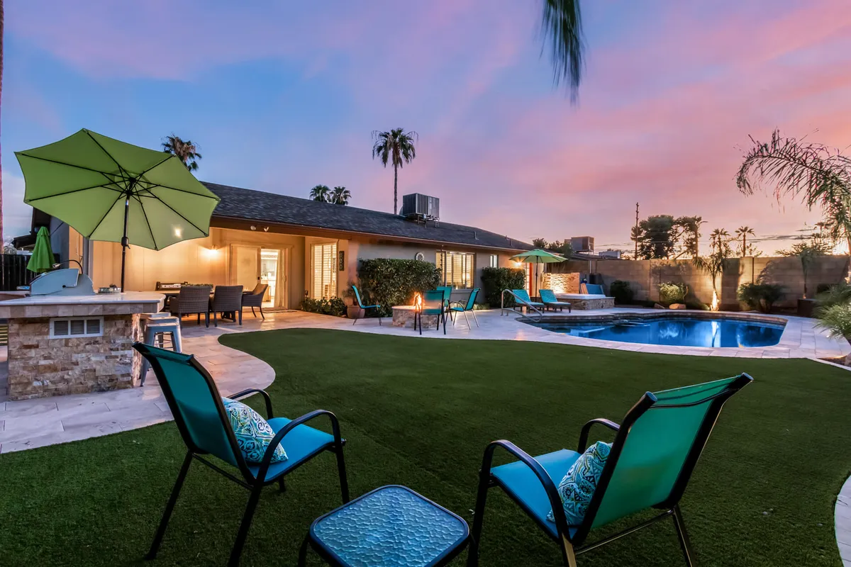 A sunset-lit outdoor area featuring a swimming pool, cozy seating, a fire pit, and a green umbrella. The inviting setup hints at post-event relaxation, socializing, and unwinding between high-energy outings.