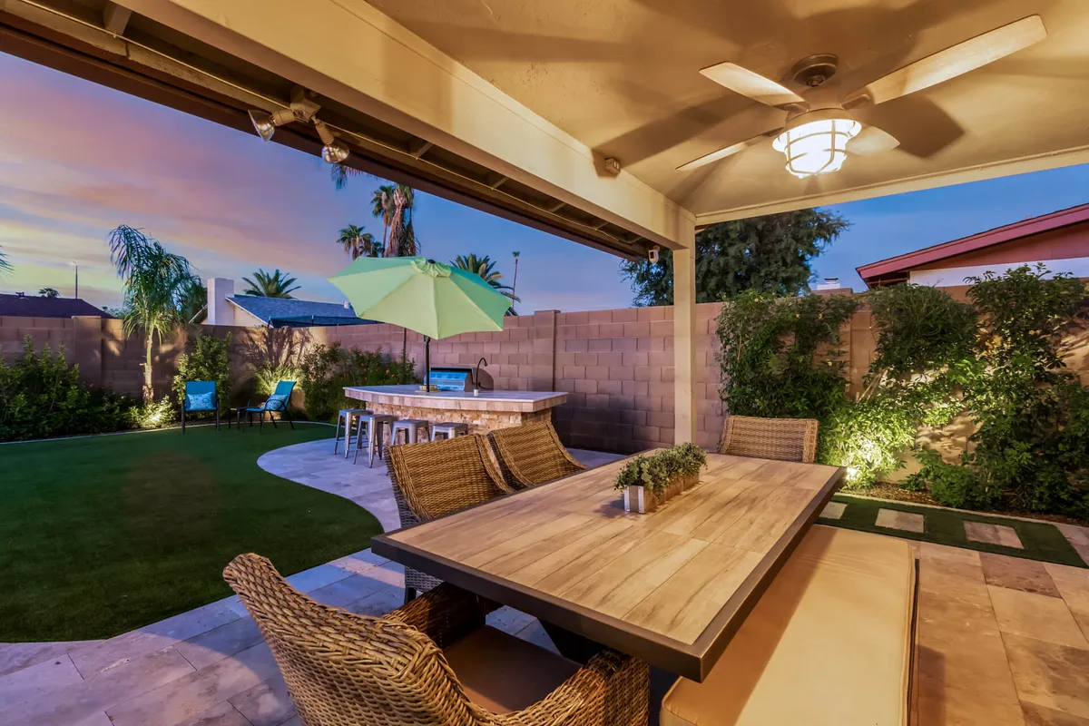 This image features an outdoor dining table and seating area shaded by an umbrella, set within a lush, landscaped garden that is dotted with palm trees. There is a barbecue area nearby, and the space is welcoming and cozy, suggesting an ideal environment for al fresco meals, social gatherings, and relaxation.