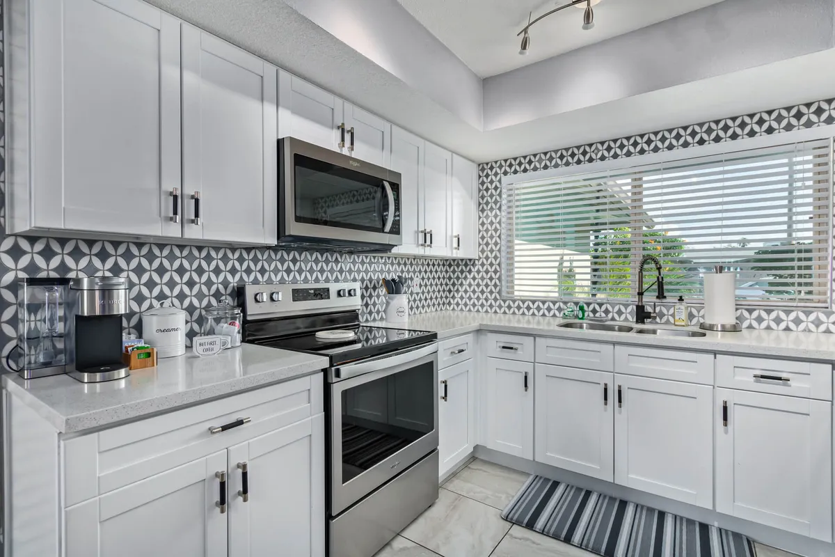 A sleek, modern kitchen featuring a striking geometric backsplash, stainless steel appliances, and a large window with blinds that allows natural light to flood the space. The countertops are spacious, ideal for meal preparation or serving. The scene suggests lively cooking, baking, and entertaining moments, perfectly matching the described 'Chef’s Corner.'